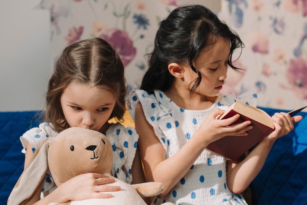 Young Girls Sitting Together at the Bed while Reading a Book and Holding a Plush Toy | Kids Car Donations