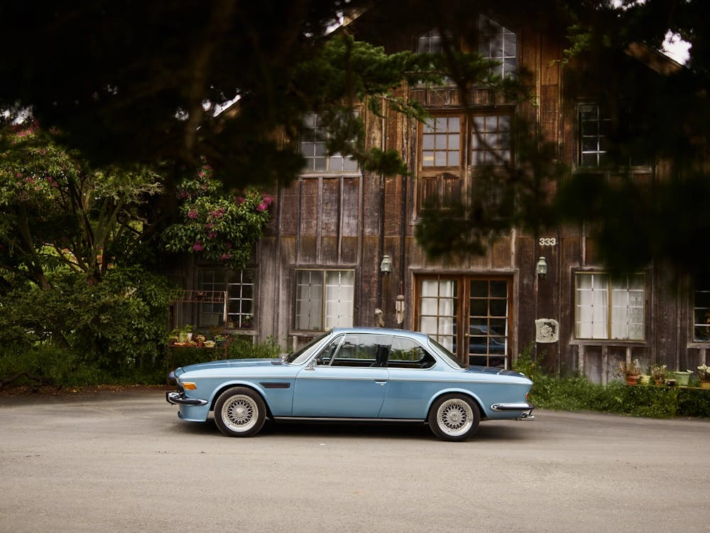 Vintage blue car parked near a rustic wooden house under large tree branches