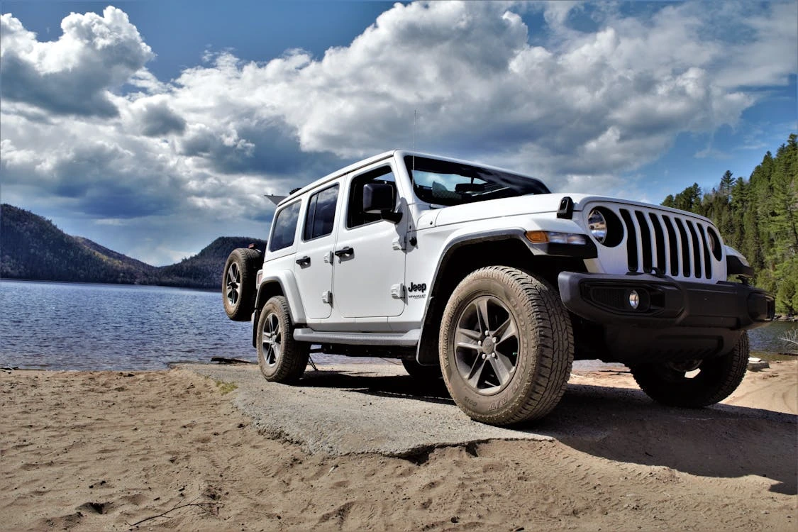 White Jeep parked on a sandy shore beside a calm lake under cloudy skies